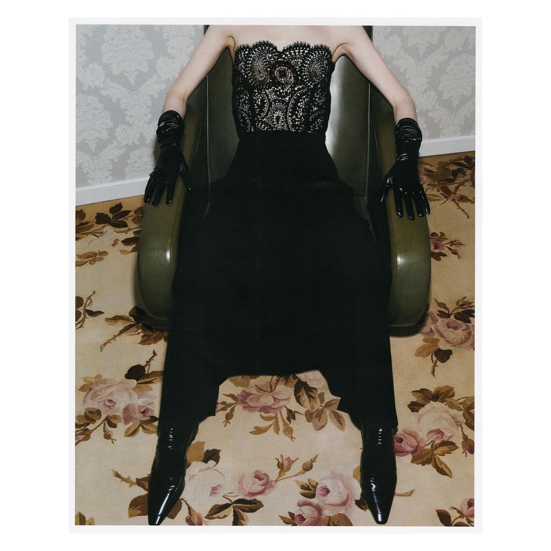Woman in black lace corset and pants sitting in vintage armchair with floral wallpaper and carpet, showcasing fashion editorial style.