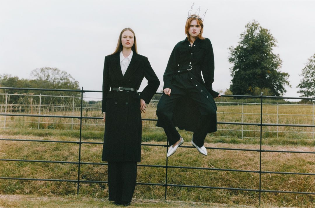 Two women in black coats pose on a rural fence line, one sitting on the fence and wearing a decorative crown, in an outdoor setting.