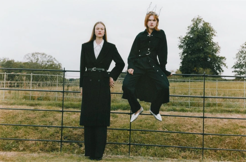 Two women in black coats pose on a rural fence line, one sitting on the fence and wearing a decorative crown, in an outdoor setting.