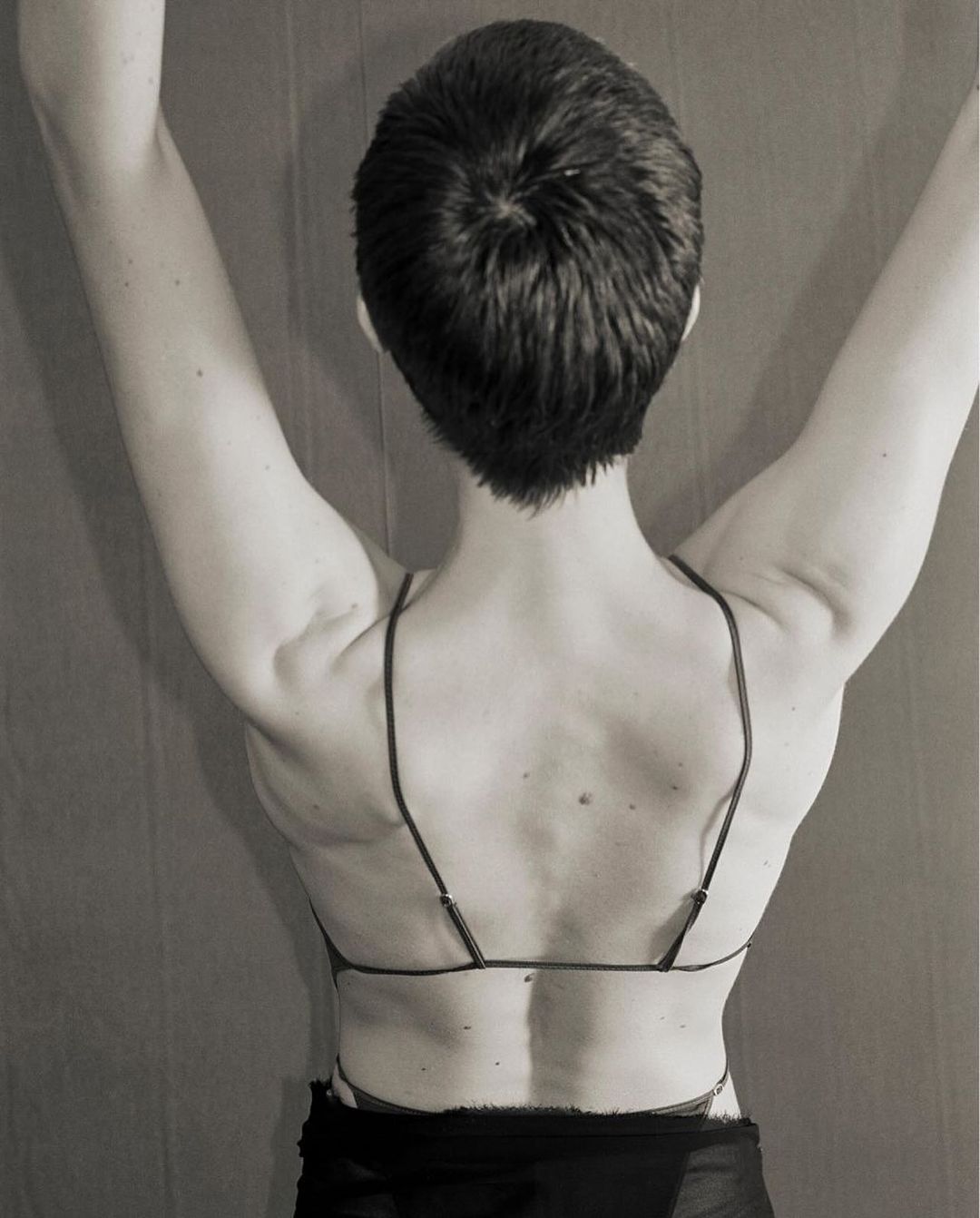 Back view of a person with short dark hair and arms raised, wearing a bra-like top with spaghetti straps against a neutral background.