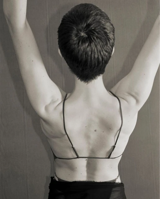 Back view of a person with short dark hair and arms raised, wearing a bra-like top with spaghetti straps against a neutral background.