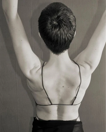 Back view of a person with short dark hair and arms raised, wearing a bra-like top with spaghetti straps against a neutral background.