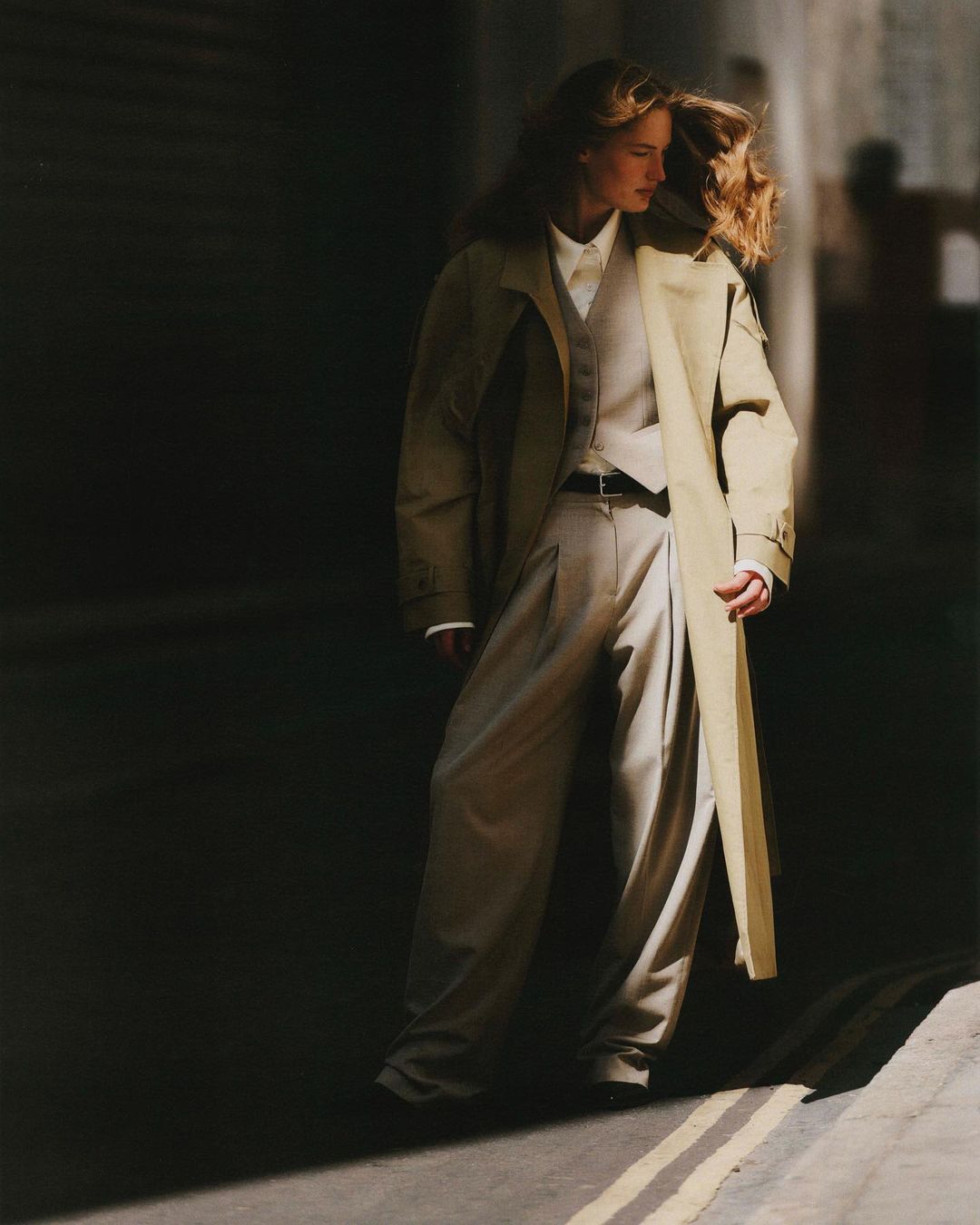 Fashion shot of a woman wearing a long coat, vest, shirt, and pants, standing on a street with a partially lit wall in the background.