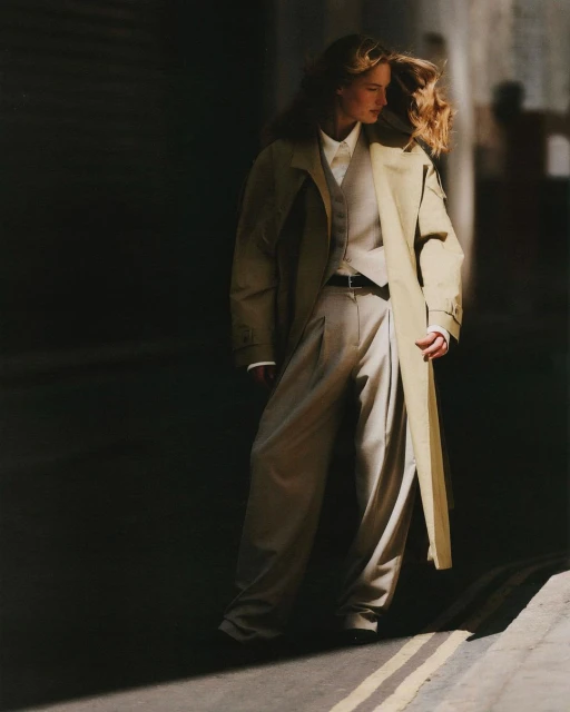 Fashion shot of a woman wearing a long coat, vest, shirt, and pants, standing on a street with a partially lit wall in the background.