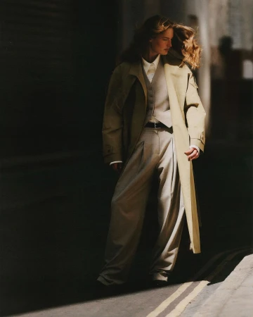Fashion shot of a woman wearing a long coat, vest, shirt, and pants, standing on a street with a partially lit wall in the background.
