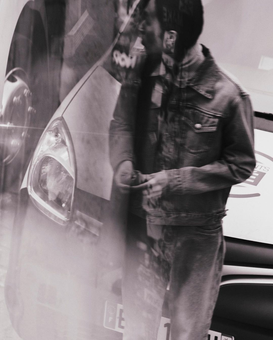 Man in a denim jacket stands near a car, with a superimposed effect creating a blended scene, rendered in black and white.