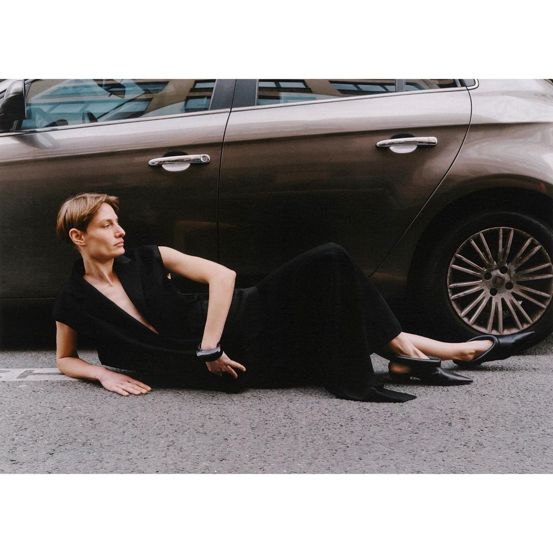 Adult woman in a black dress lying on the asphalt road next to a metallic brown car in an urban outdoor setting.
