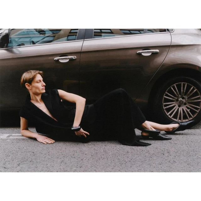 Adult woman in a black dress lying on the asphalt road next to a metallic brown car in an urban outdoor setting.