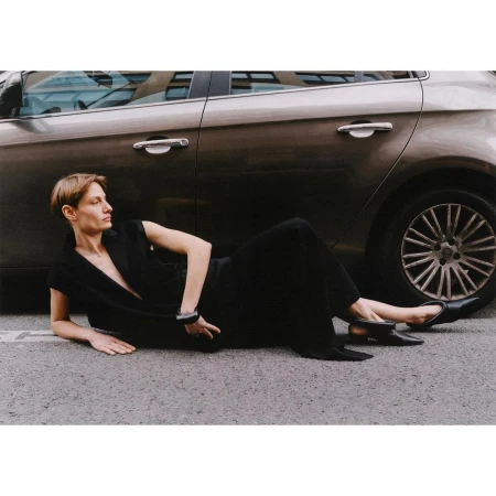 Adult woman in a black dress lying on the asphalt road next to a metallic brown car in an urban outdoor setting.
