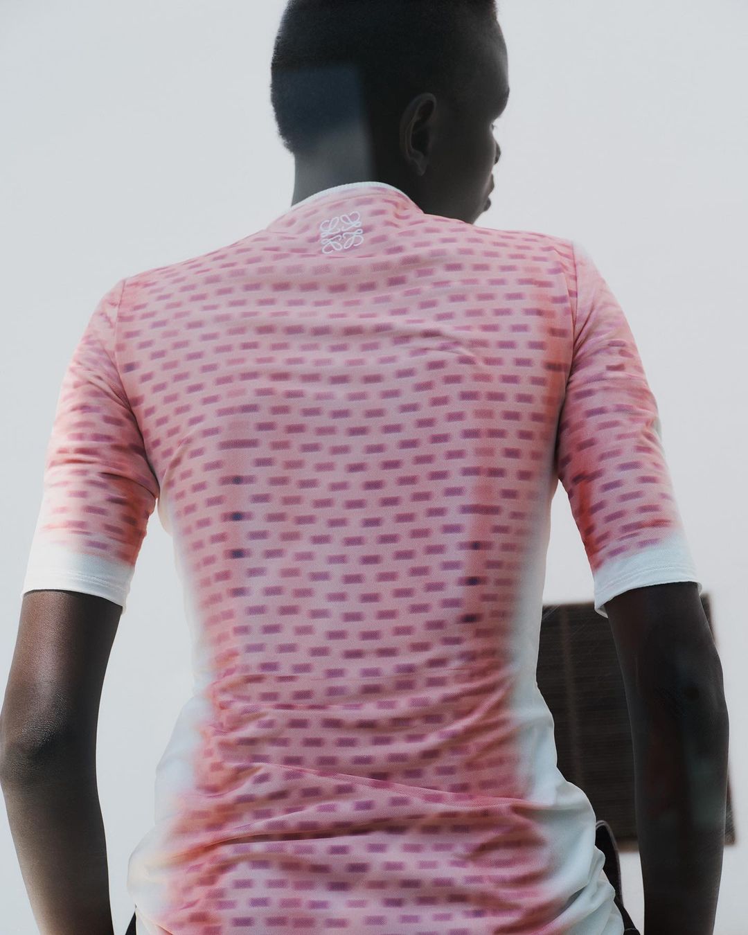Back view of an adult wearing a pink short-sleeved shirt with a geometric pattern, standing in front of a light neutral wall.