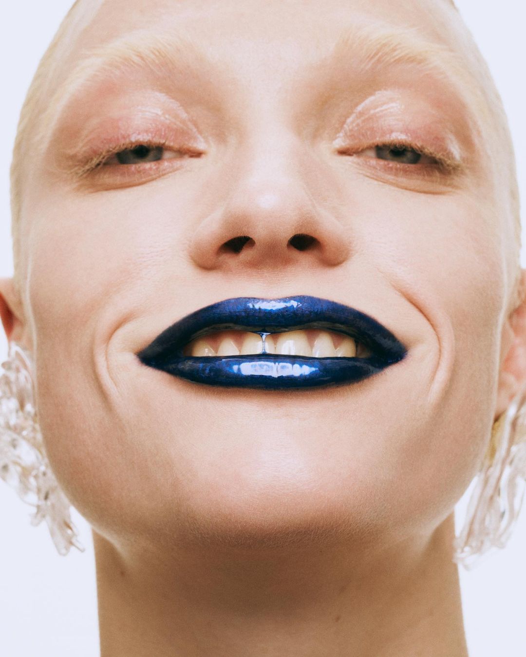 Close-up beauty shot of a woman with pale skin and dark blue glossy lipstick smiling, wearing clear earrings against a white background.