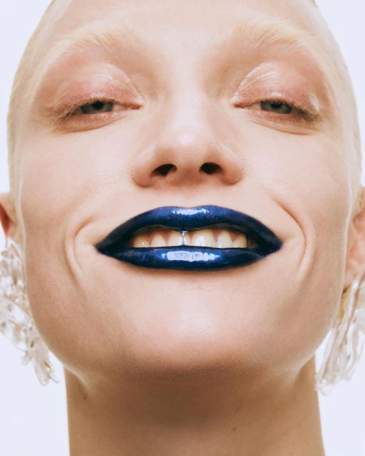 Close-up beauty shot of a woman with pale skin and dark blue glossy lipstick smiling, wearing clear earrings against a white background.
