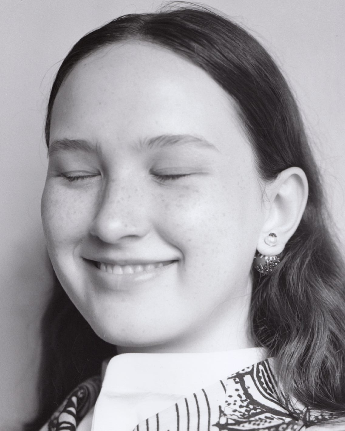 Close-up portrait of a young woman with closed eyes and a serene expression, wearing patterned clothing and earrings against a plain background.