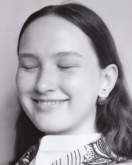 Close-up portrait of a young woman with closed eyes and a serene expression, wearing patterned clothing and earrings against a plain background.
