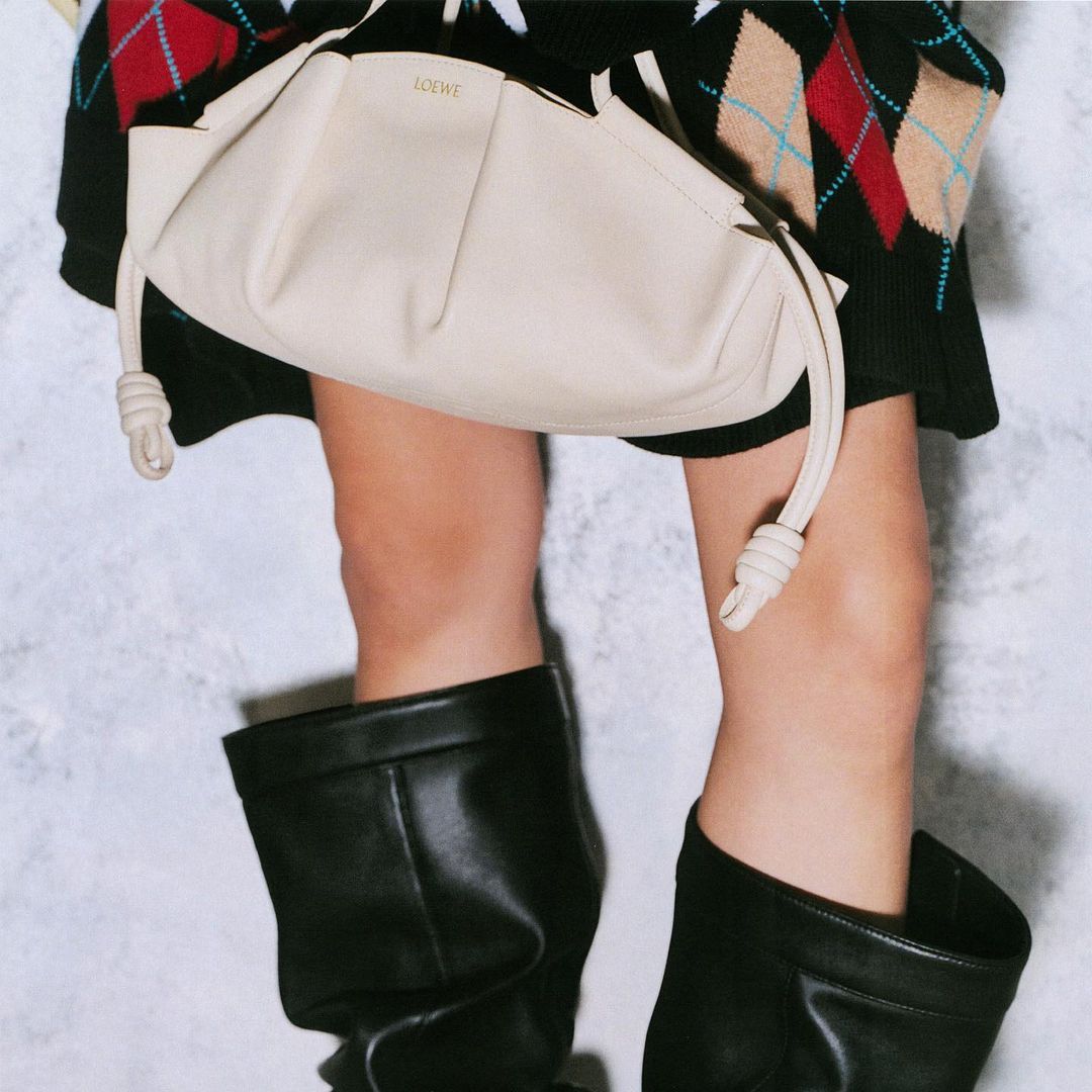 Close-up of a woman in black leather boots with a cream-colored Loewe bag, showcasing fashion and style against a textured background.