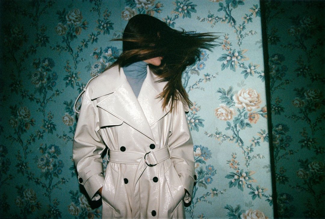 A woman in a tan trench coat stands against a teal floral wallpaper background with her hair blowing across her face.