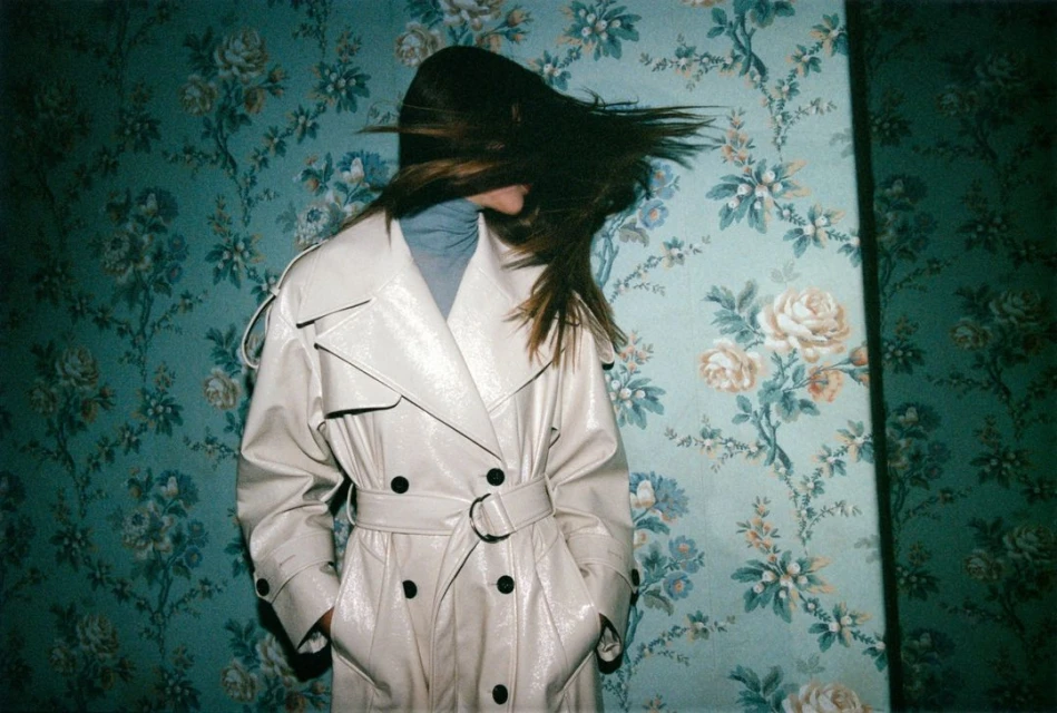 A woman in a tan trench coat stands against a teal floral wallpaper background with her hair blowing across her face.