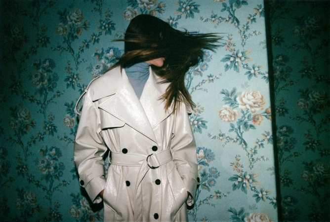 A woman in a tan trench coat stands against a teal floral wallpaper background with her hair blowing across her face.