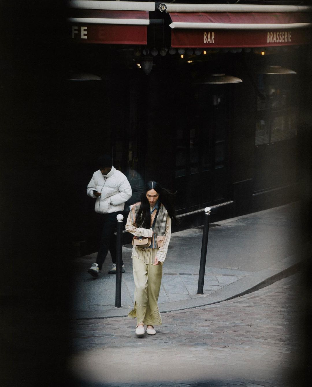 Woman walks on a cobblestone street in front of a cafe with another person in the background wearing a puffer jacket in urban setting.