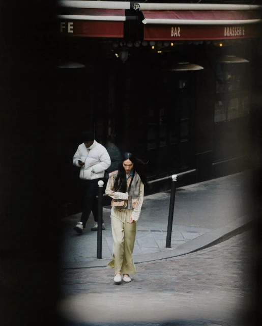 Woman walks on a cobblestone street in front of a cafe with another person in the background wearing a puffer jacket in urban setting.