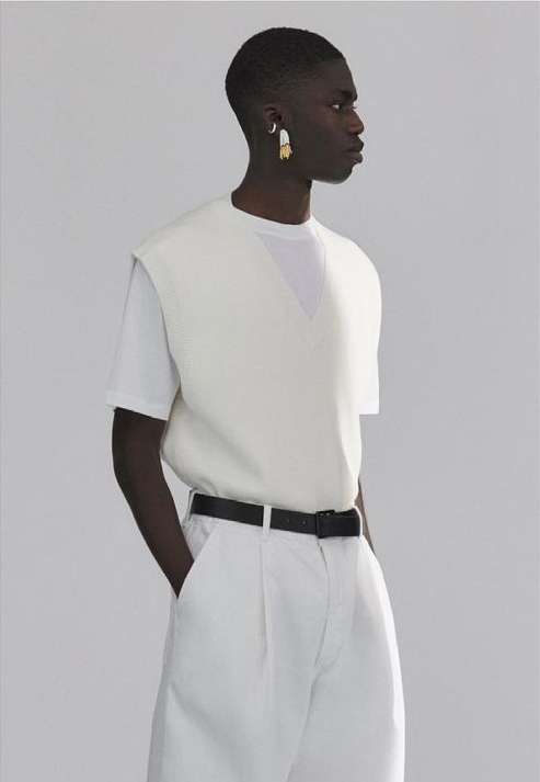 Dark-skinned man in a white sleeveless sweater and pants poses in a photo studio with a black belt and white and gold earrings.