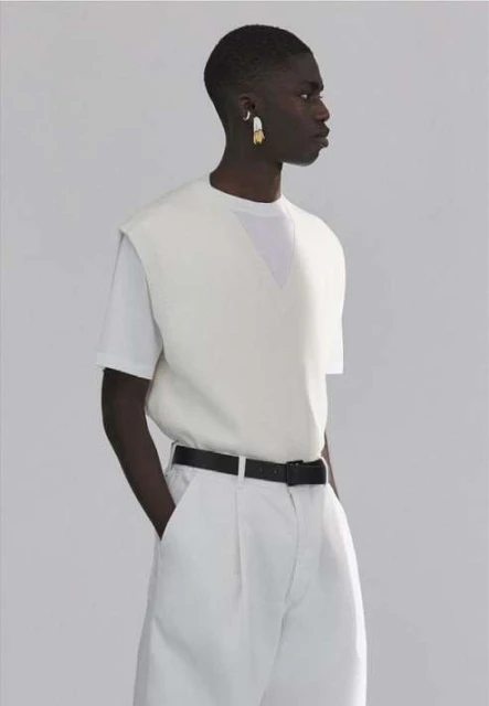 Dark-skinned man in a white sleeveless sweater and pants poses in a photo studio with a black belt and white and gold earrings.