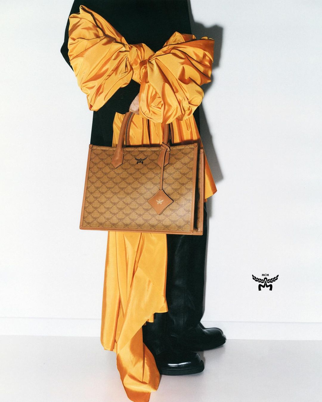 Fashion-forward shot of a handbag with repeating patterns, paired with an oversized gold bow and black apparel against a white backdrop.