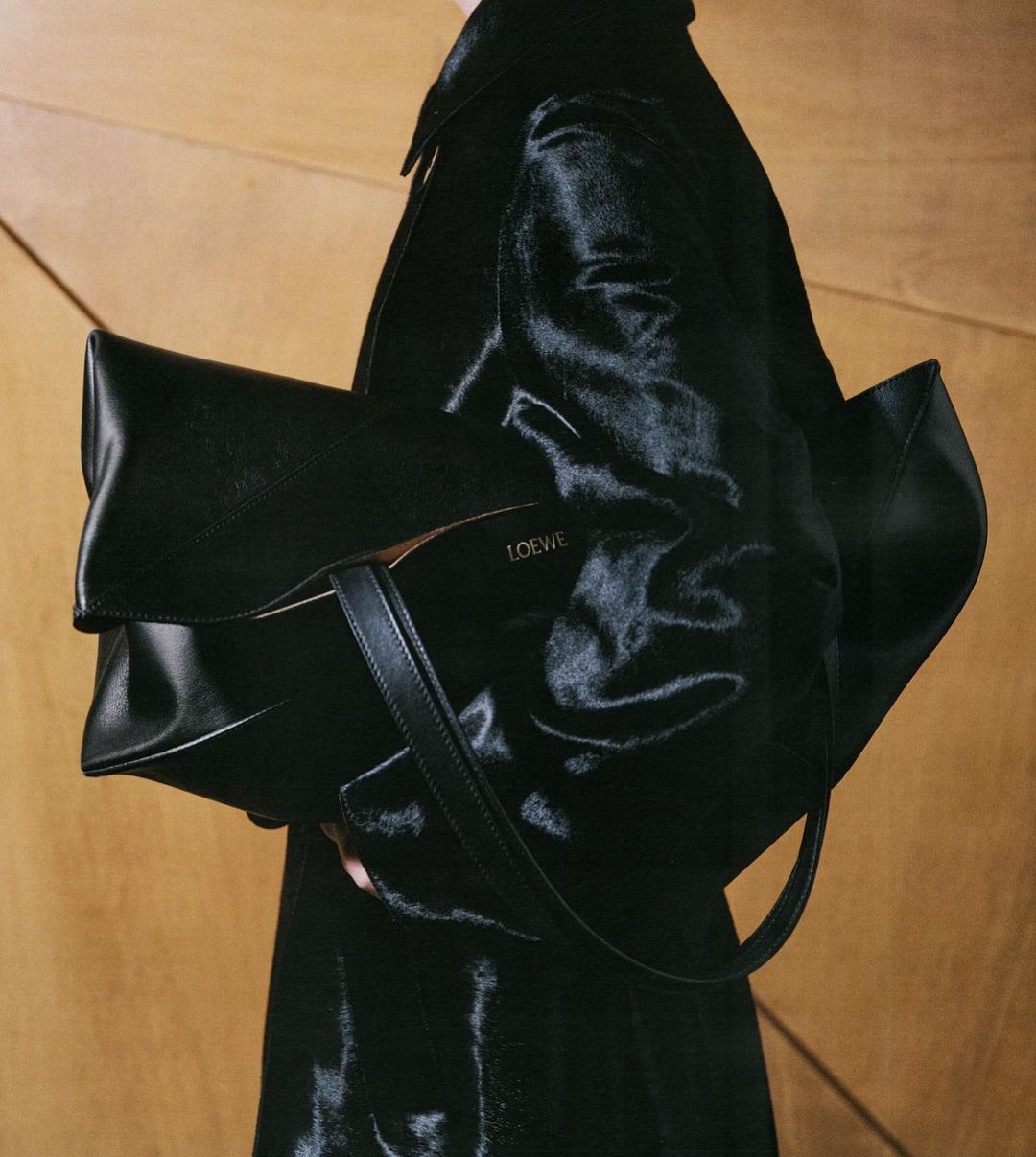 Black leather handbag with the Loewe logo, styled with a dark coat against a paneled wood backdrop in a studio fashion shot.