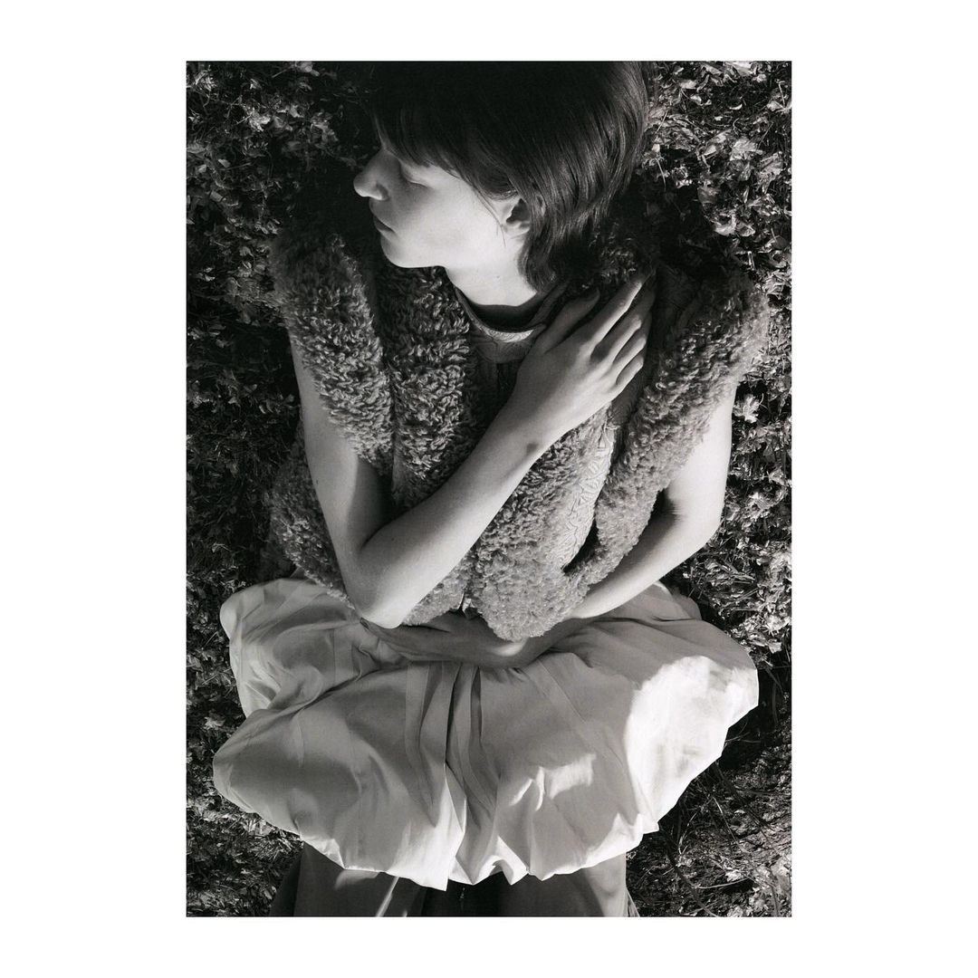 Monochrome image of a young woman lying in a field, wearing a furry vest and a billowing skirt. She has her eyes closed and arms crossed.