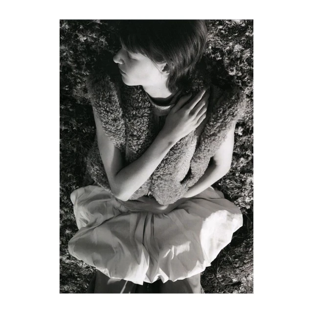 Monochrome image of a young woman lying in a field, wearing a furry vest and a billowing skirt. She has her eyes closed and arms crossed.