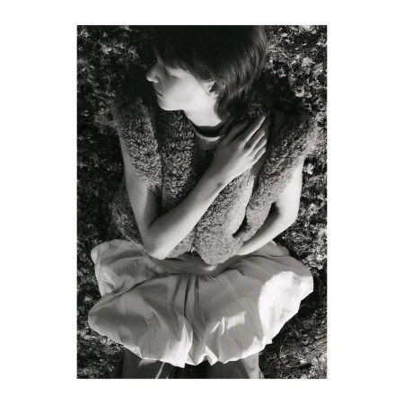 Monochrome image of a young woman lying in a field, wearing a furry vest and a billowing skirt. She has her eyes closed and arms crossed.