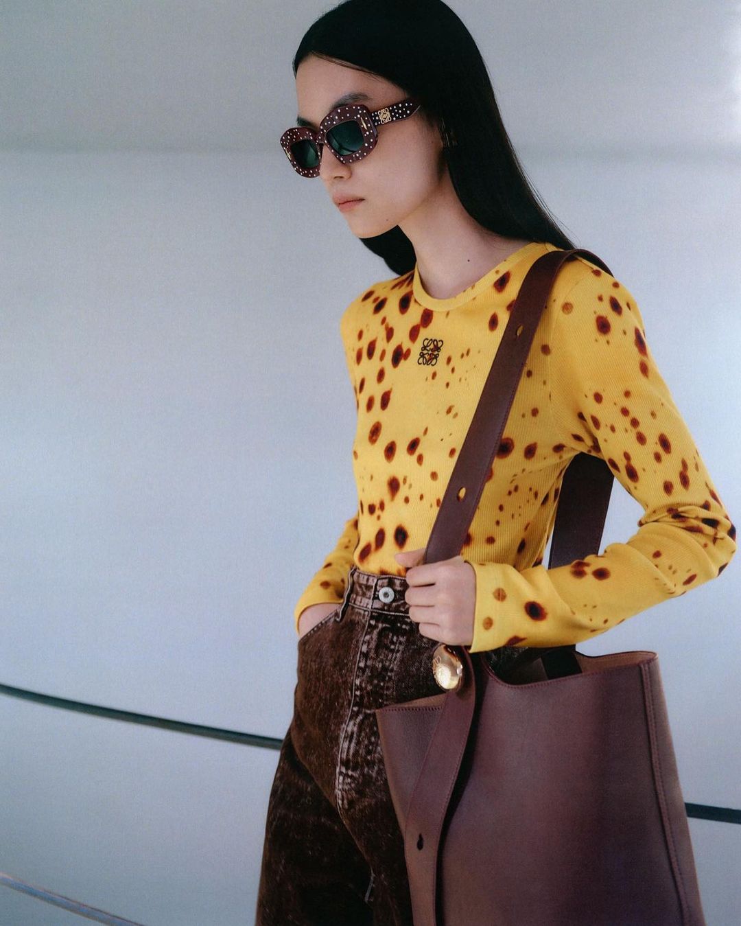 Elegant woman in yellow long-sleeve top and brown jeans, carrying a brown leather tote bag and wearing sunglasses in a studio setting.