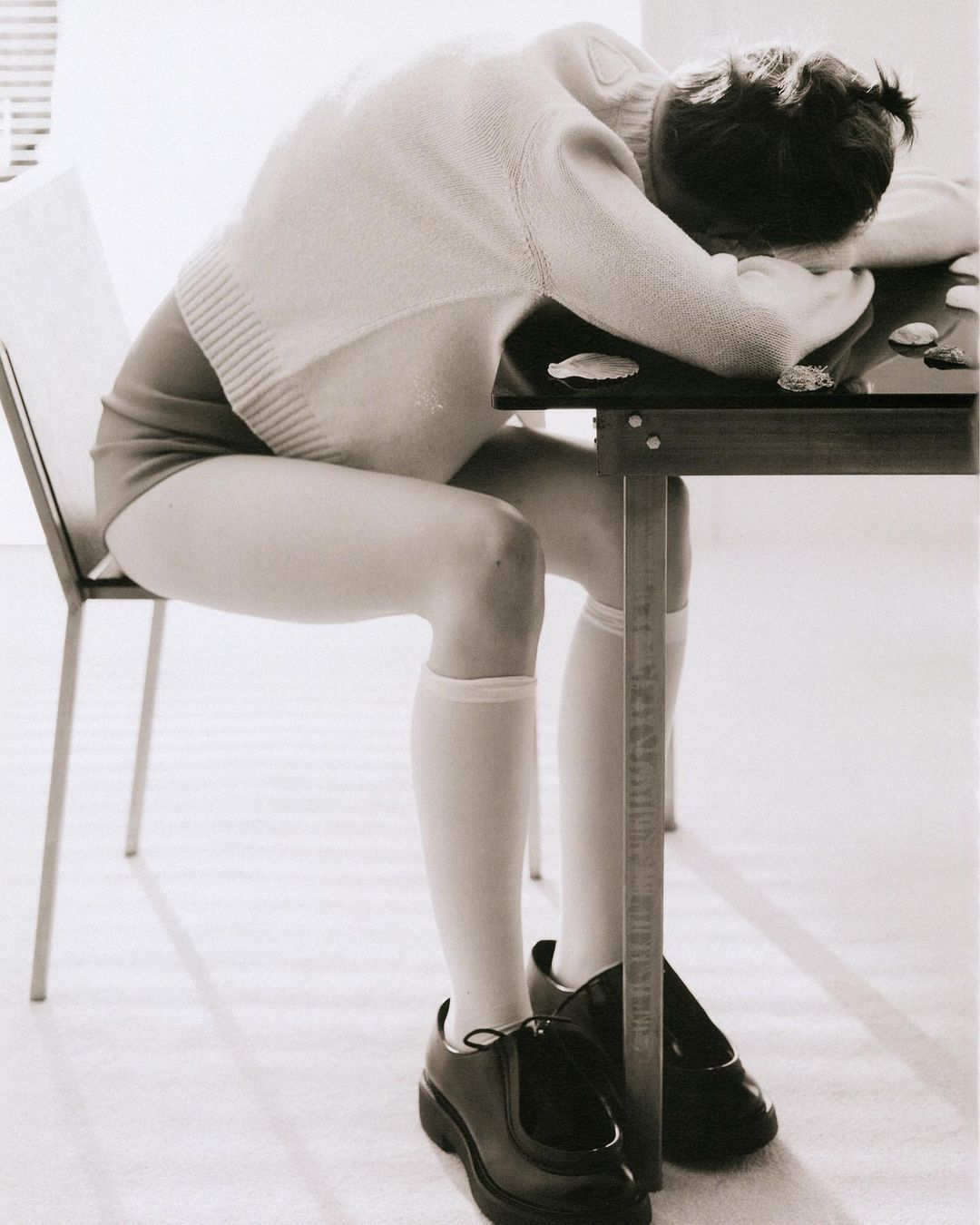 Person seated at table with head down, wearing a sweater, leotard, knee socks, and shoes. Black and white photo, indoor setting.