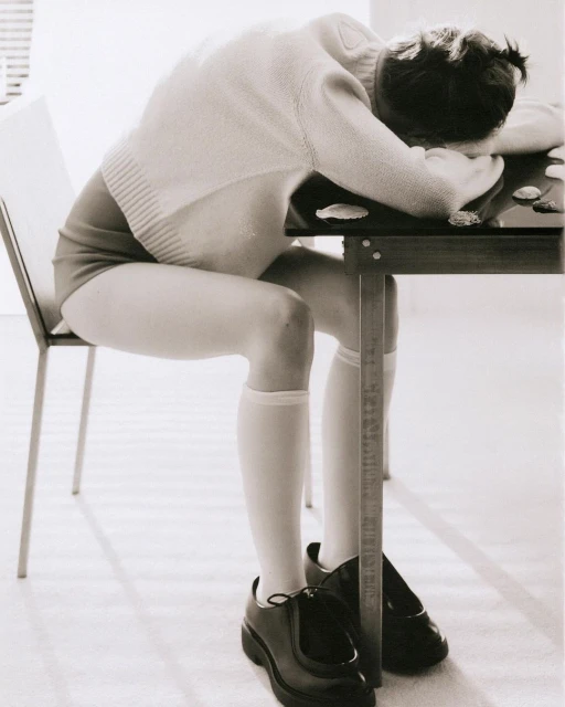Person seated at table with head down, wearing a sweater, leotard, knee socks, and shoes. Black and white photo, indoor setting.