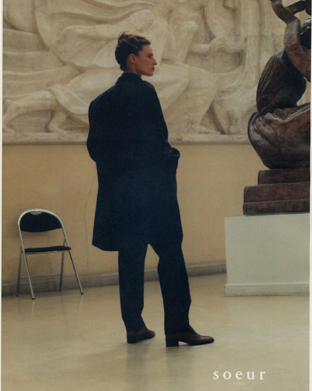 Woman in long dark coat standing in an art gallery with sculptures and a chair. Fashion editorial-style photograph with vintage aesthetic.
