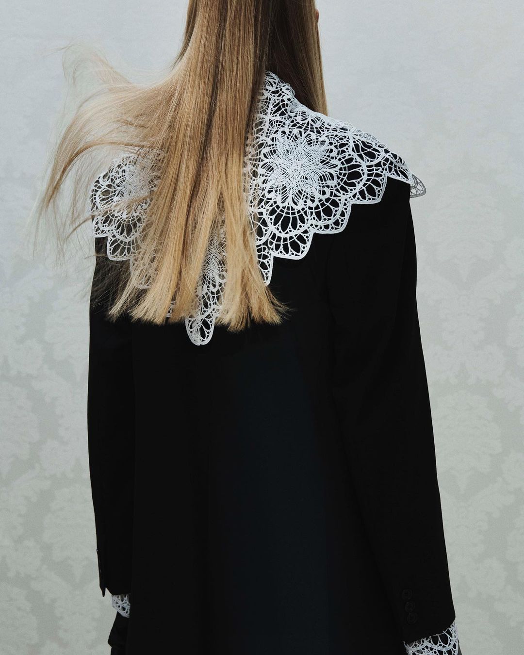 Blonde woman's back, wearing black dress with large white lace collar, against neutral background. Fashion photography. 