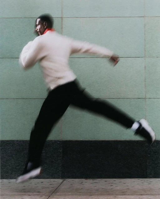 Motion-blurred image of an adult man running on a sidewalk in a cream-colored sweatshirt and black pants against a textured wall.