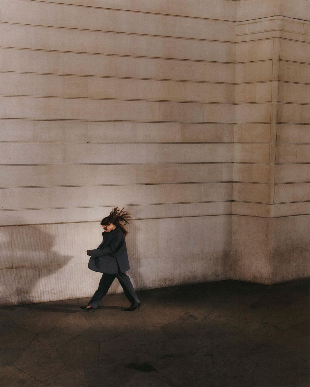 Woman in a dark suit walks in front of a textured tan wall in an urban environment, capturing a moment of city life with long shadows.