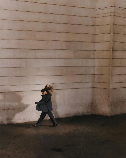 Woman in a dark suit walks in front of a textured tan wall in an urban environment, capturing a moment of city life with long shadows.