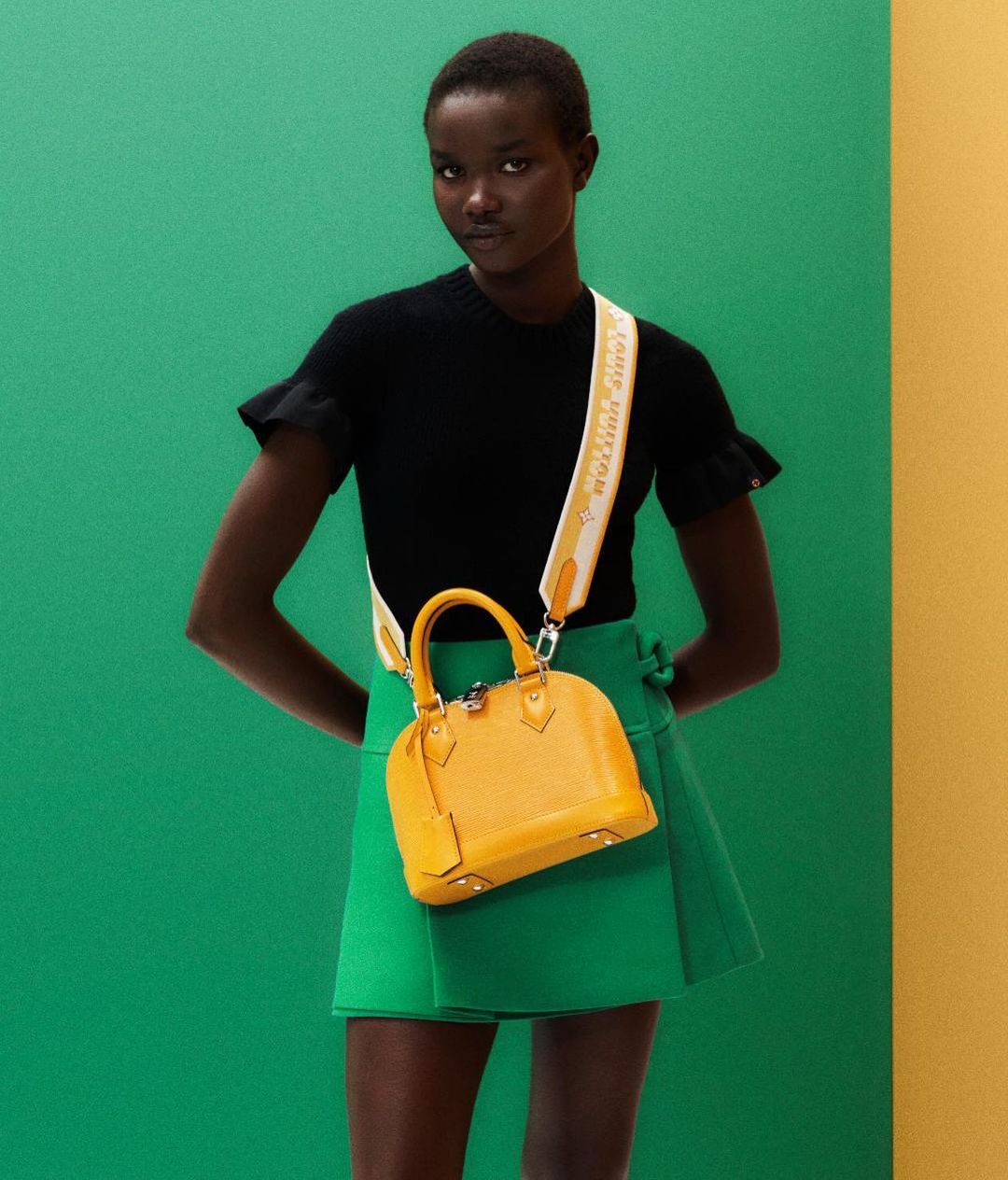 A woman poses in a black top and green skirt with a yellow handbag against a two-tone green and yellow background in a studio shot.