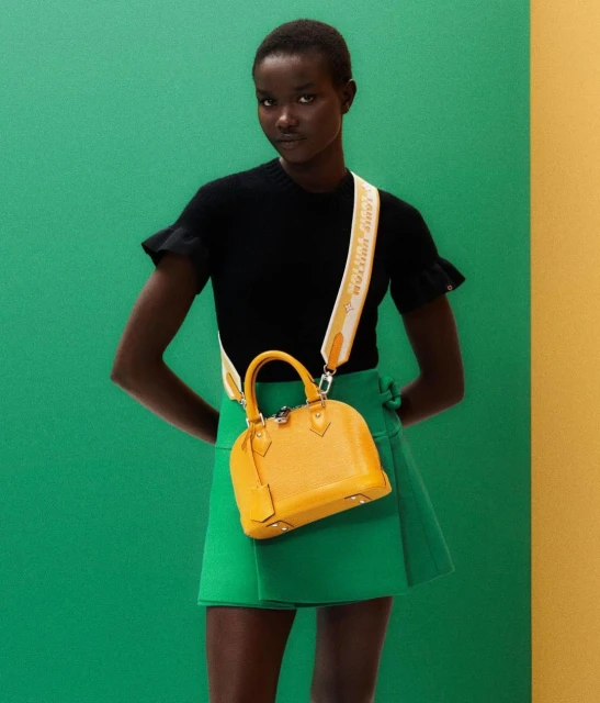 A woman poses in a black top and green skirt with a yellow handbag against a two-tone green and yellow background in a studio shot.