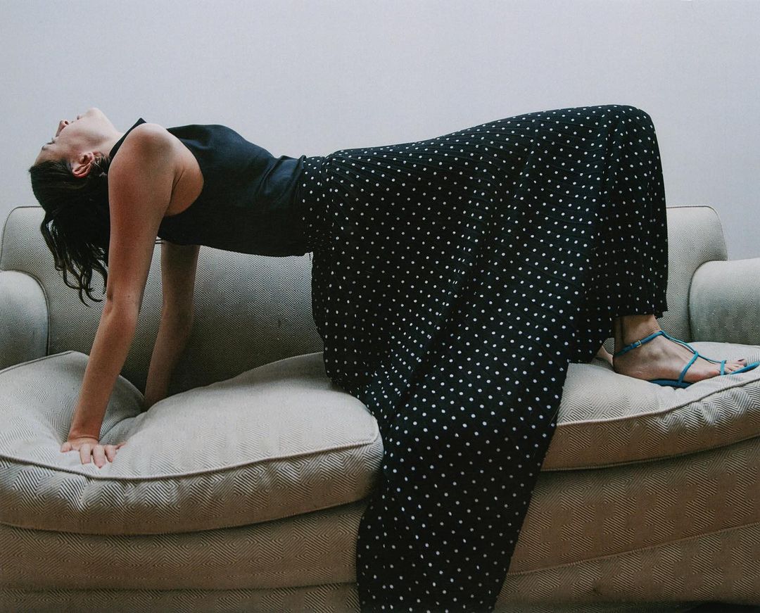 Woman in a black top and long black and white polka dot skirt arches back on a light beige sofa in a brightly lit interior.