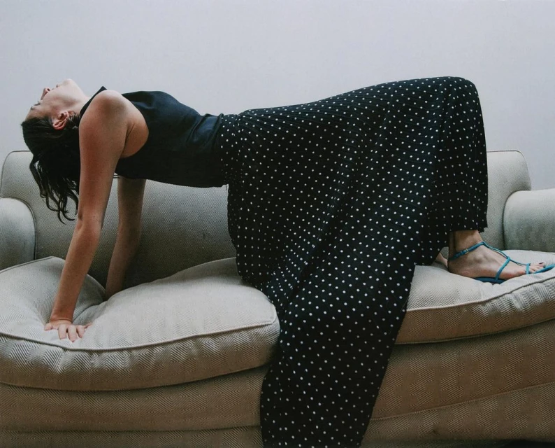 Woman in a black top and long black and white polka dot skirt arches back on a light beige sofa in a brightly lit interior.