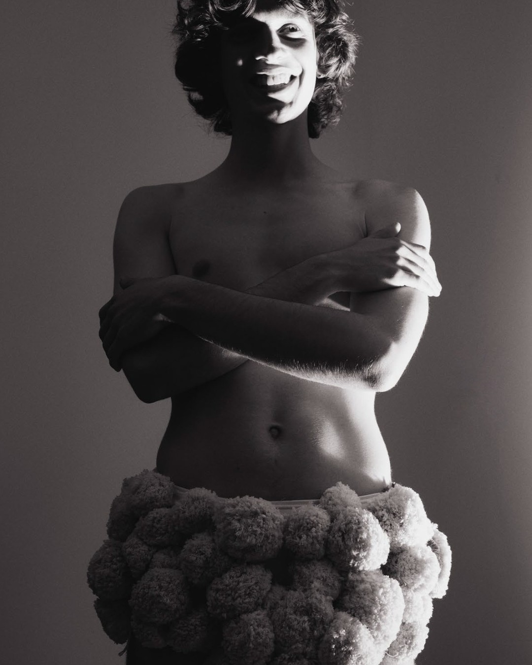 Black and white portrait of a smiling, partially shadowed person with curly hair, wearing a fluffy skirt, against a simple background.