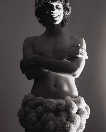 Black and white portrait of a smiling, partially shadowed person with curly hair, wearing a fluffy skirt, against a simple background.