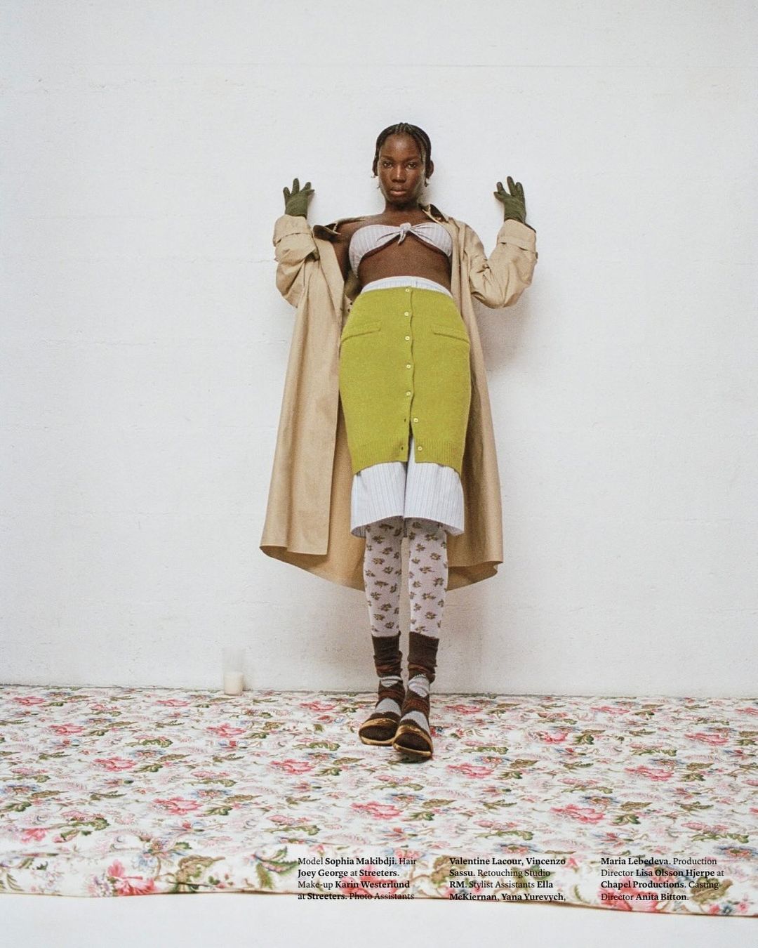 Fashionable Black woman in a tan trench coat and knitwear poses in front of a white wall on a floral patterned surface in a studio.