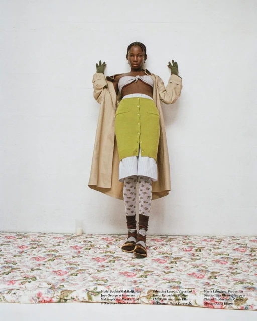 Fashionable Black woman in a tan trench coat and knitwear poses in front of a white wall on a floral patterned surface in a studio.