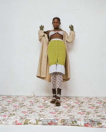 Fashionable Black woman in a tan trench coat and knitwear poses in front of a white wall on a floral patterned surface in a studio.