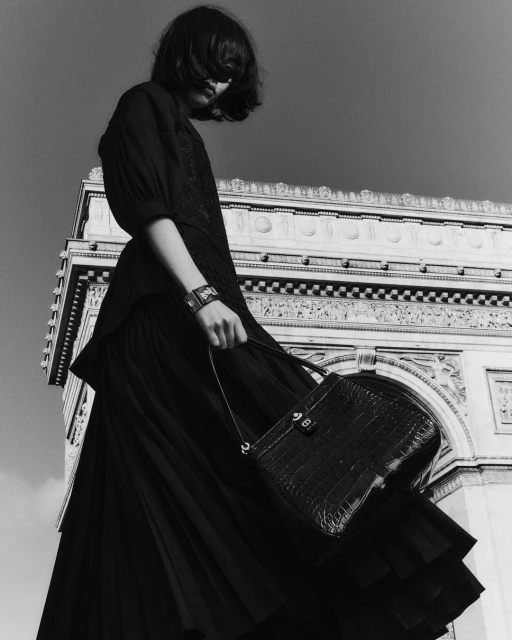 Fashion shot of a woman in a dark dress holding a bag, standing in front of the Arc de Triomphe in Paris, France. Low angle, black and white.