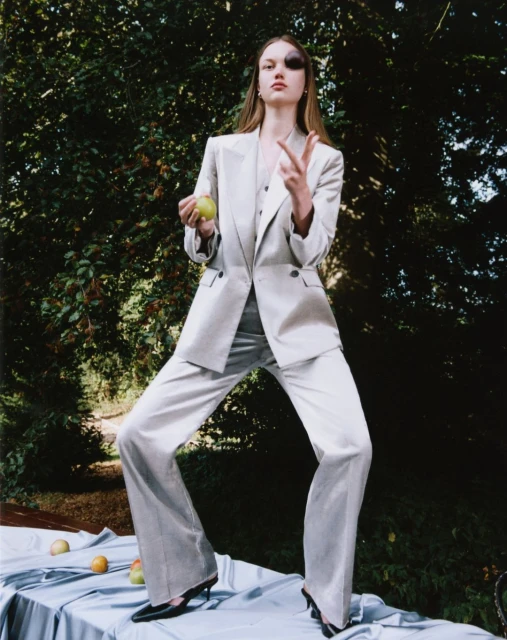 Fashion model in a light grey pantsuit stands on a table in a garden setting, holding an apple and gesturing with her hand.